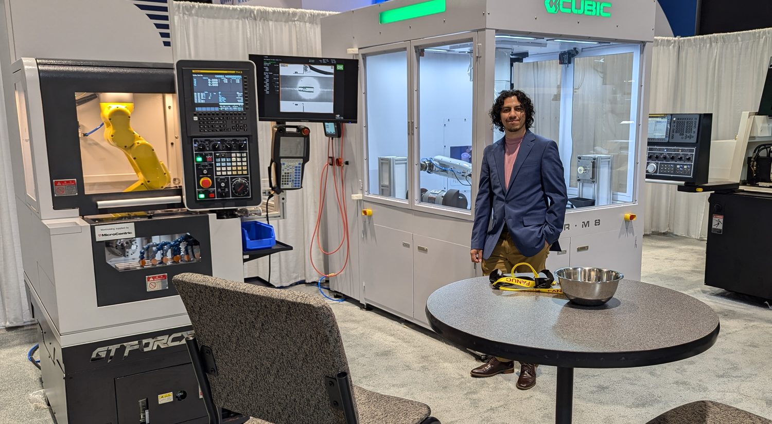 David Facio standing between a gang-tool CNC lathe with robot loader and a robotic deburring cell at the Cubic Machinery exhibit, IMTS 2024
