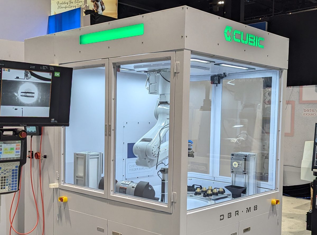 Fully guarded robotic deburring cell with glass panels and a green status light bar on a trade show floor