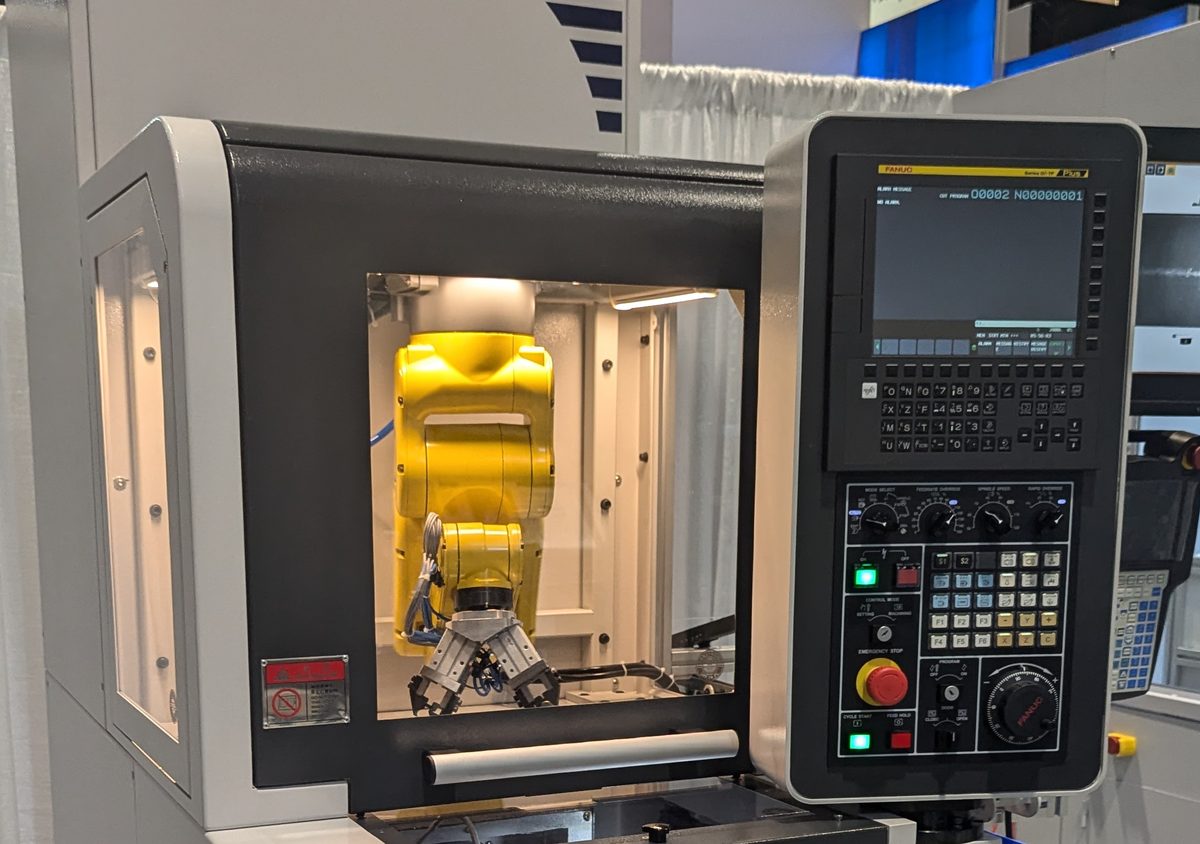 FANUC robot mounted inside a guarded loader enclosure above a gang-tool CNC lathe, next to the FANUC CNC control panel