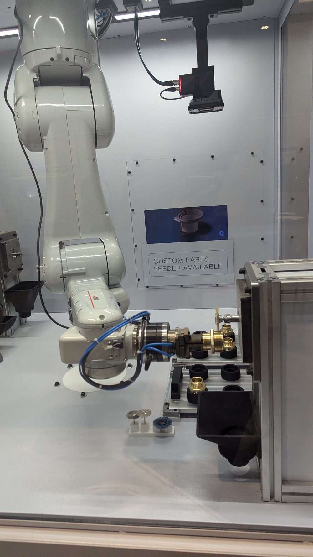 Six-axis robot inside the deburring cell holding a brass fitting against a finishing station, with a parts nest and overhead machine-vision camera