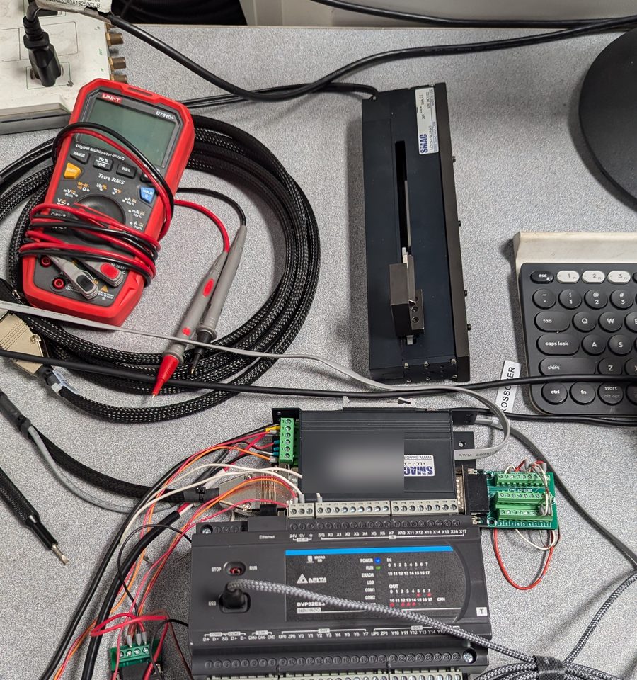 Bench test setup with a SMAC linear voice-coil actuator, its controller, a Delta PLC, a terminal breakout board and a digital multimeter